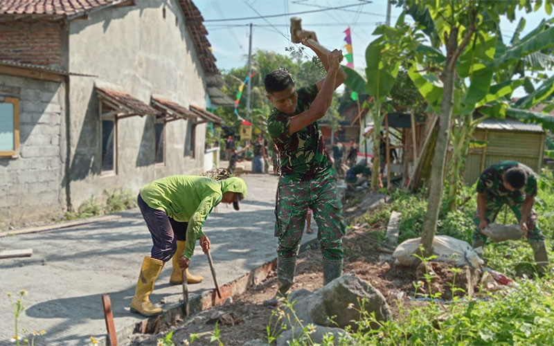 Menggunakan palu godem, Satgas TMMD memecahkan batu besar. Pecahan batu itu, selanjutnya digunakan untuk melapisi bagian bawah pengecoran jalan yang berlumpur, agar konstruksi jalan lebih kuat.